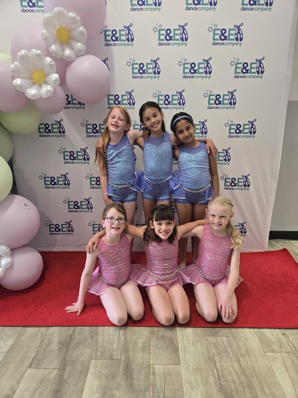 Six young dancers in pink and purple costumes posing together at recital dress rehearsal.