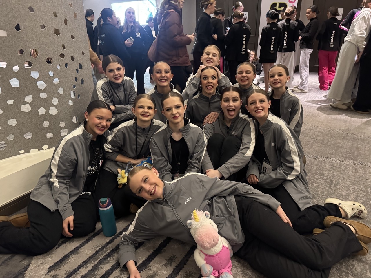 E&E Dance Company dancers in matching gray jackets smiling and posing together in a hallway.