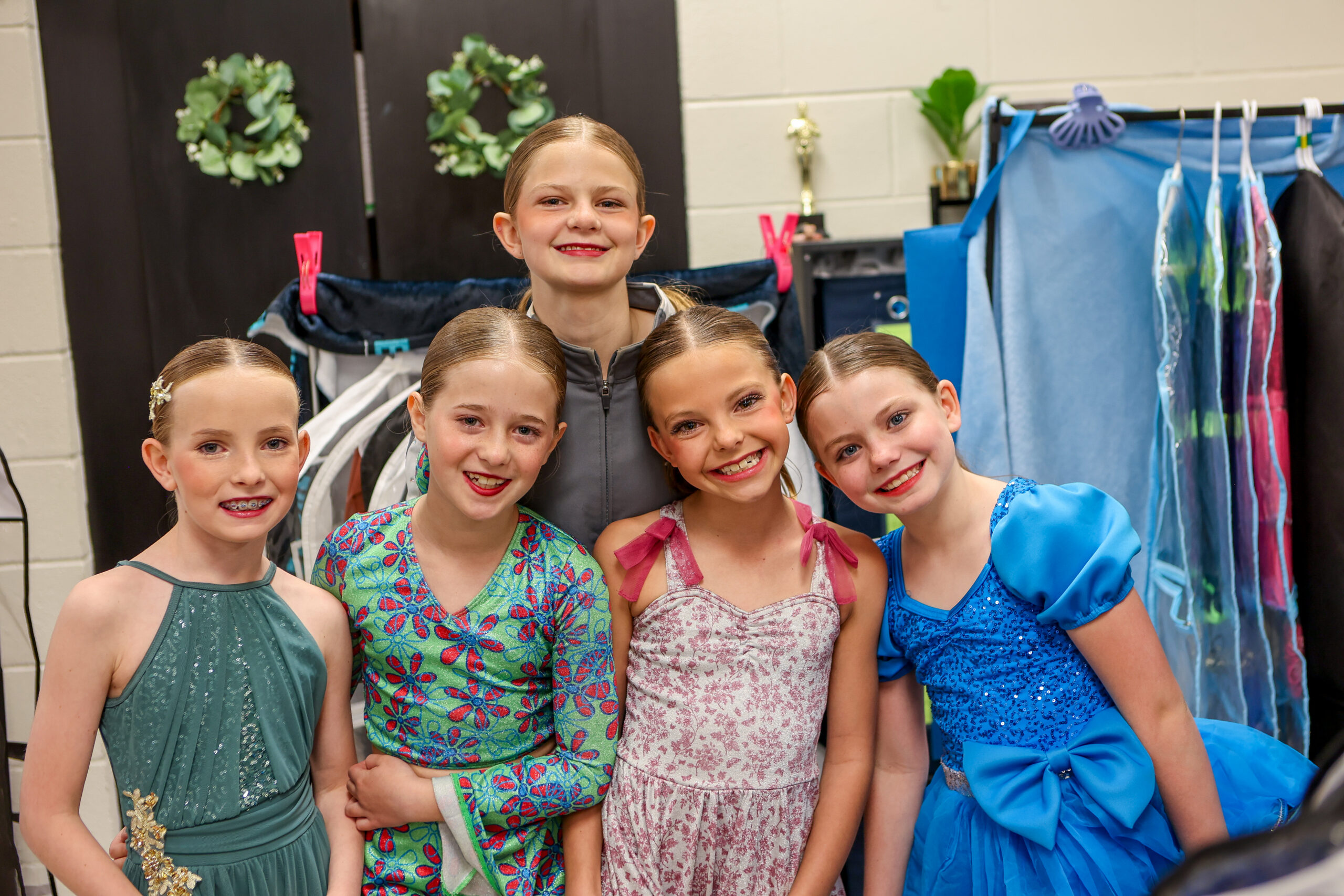 Five E&E Dance Company dancers smiling together backstage in colorful showcase costumes and a gray team jacket.