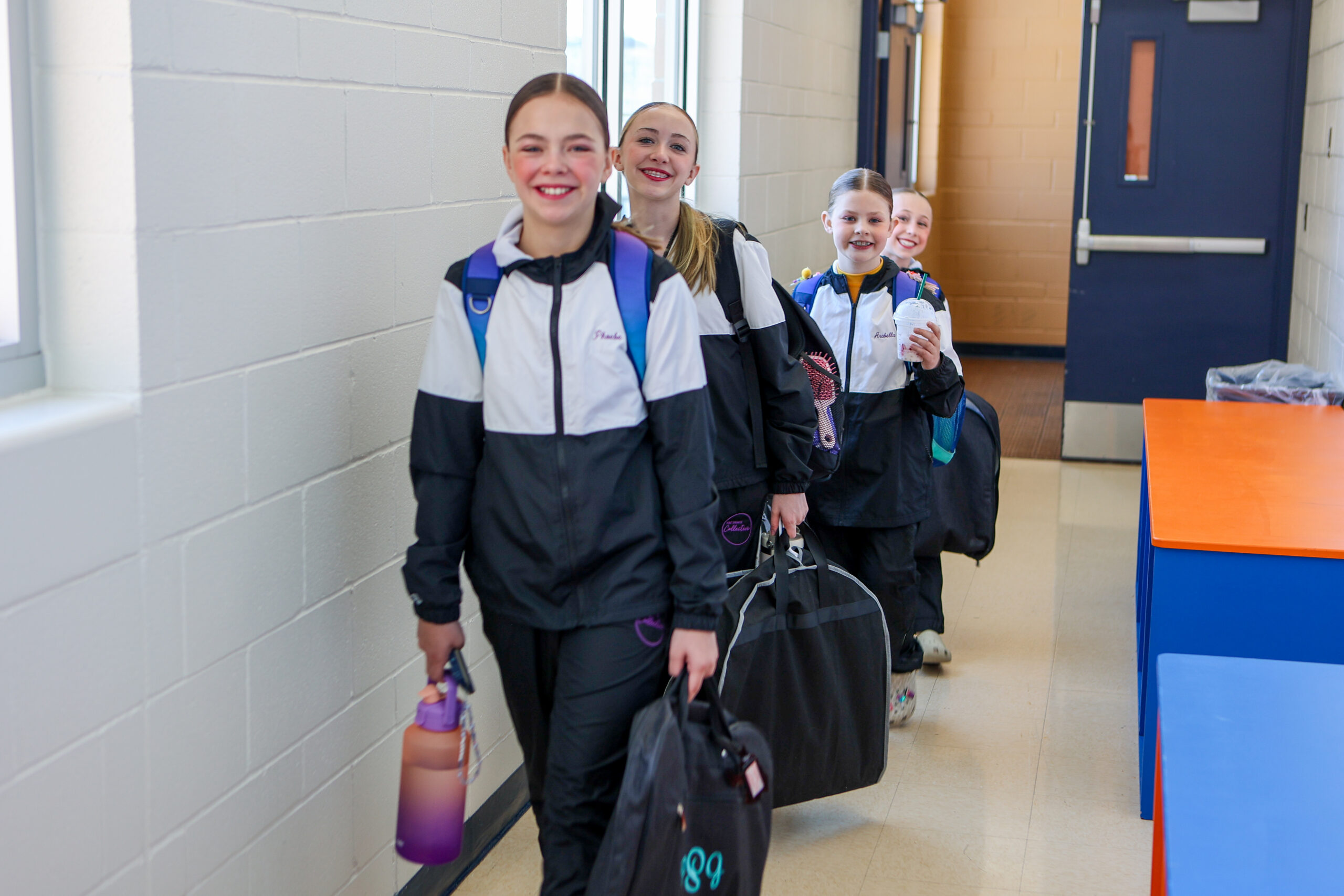 Four E&E Dance Company dancers in black and white team jackets carrying dance bags in a hallway.