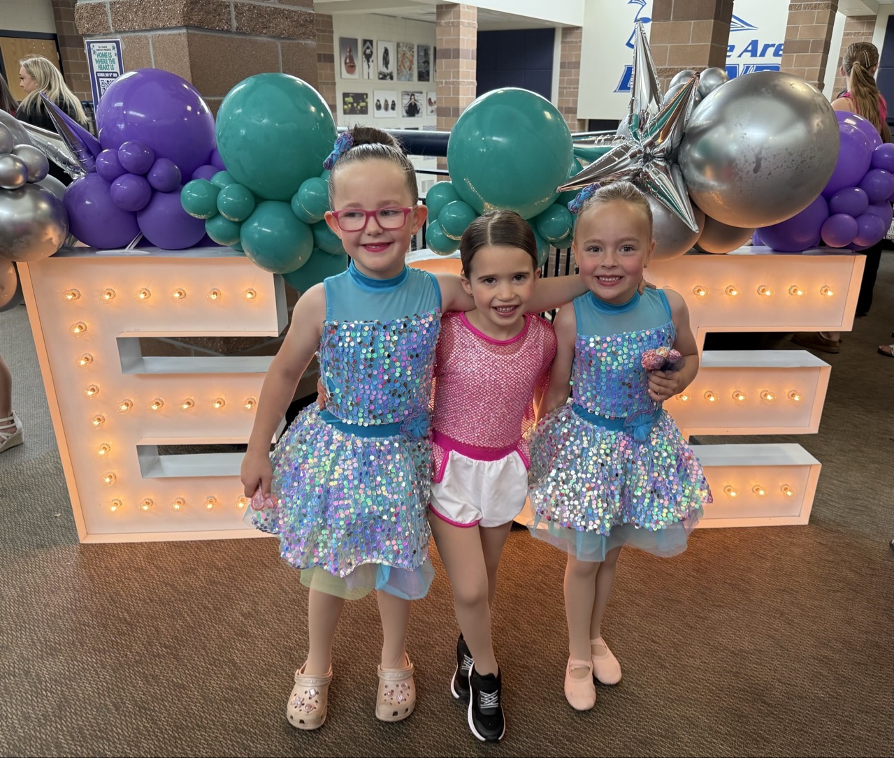 Three young E&E Dance Company dancers in sparkling blue and pink recital costumes posing together beside balloons and illuminated letters.