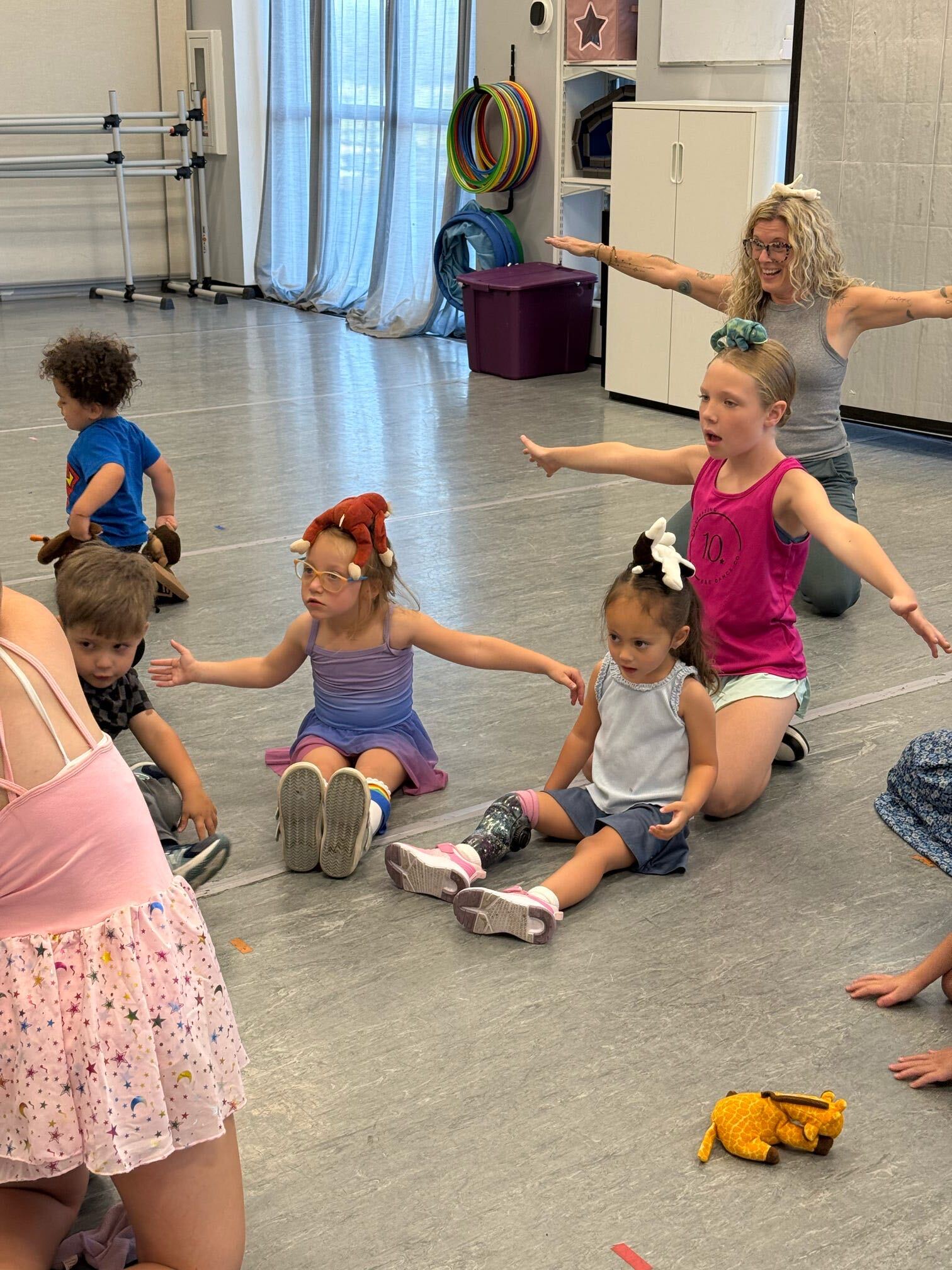 A teacher and young dancers practicing seated movements with their arms extended in the dance studio.