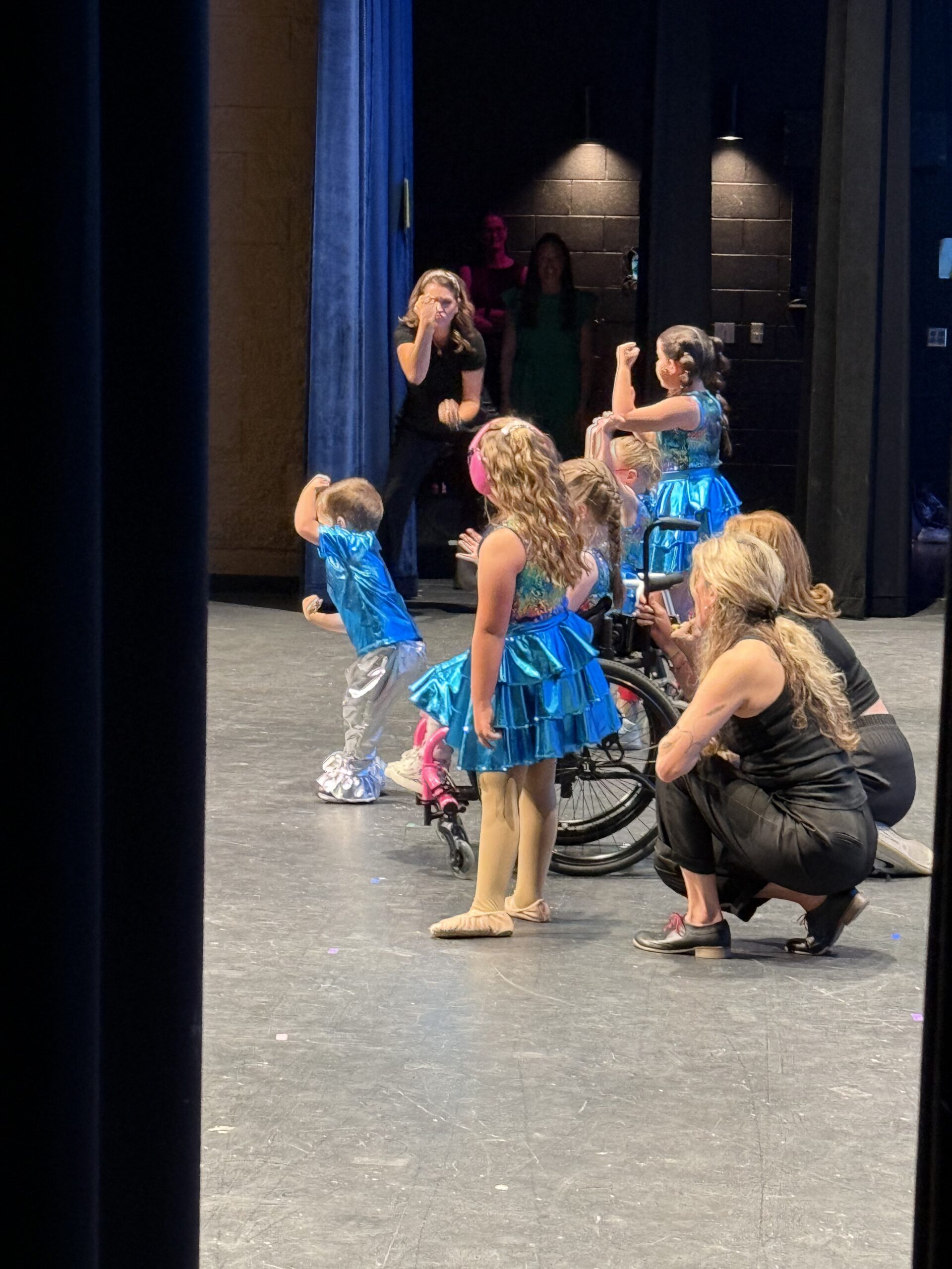Dancspiration dancers in turquoise costumes performing on stage with support from their teachers.