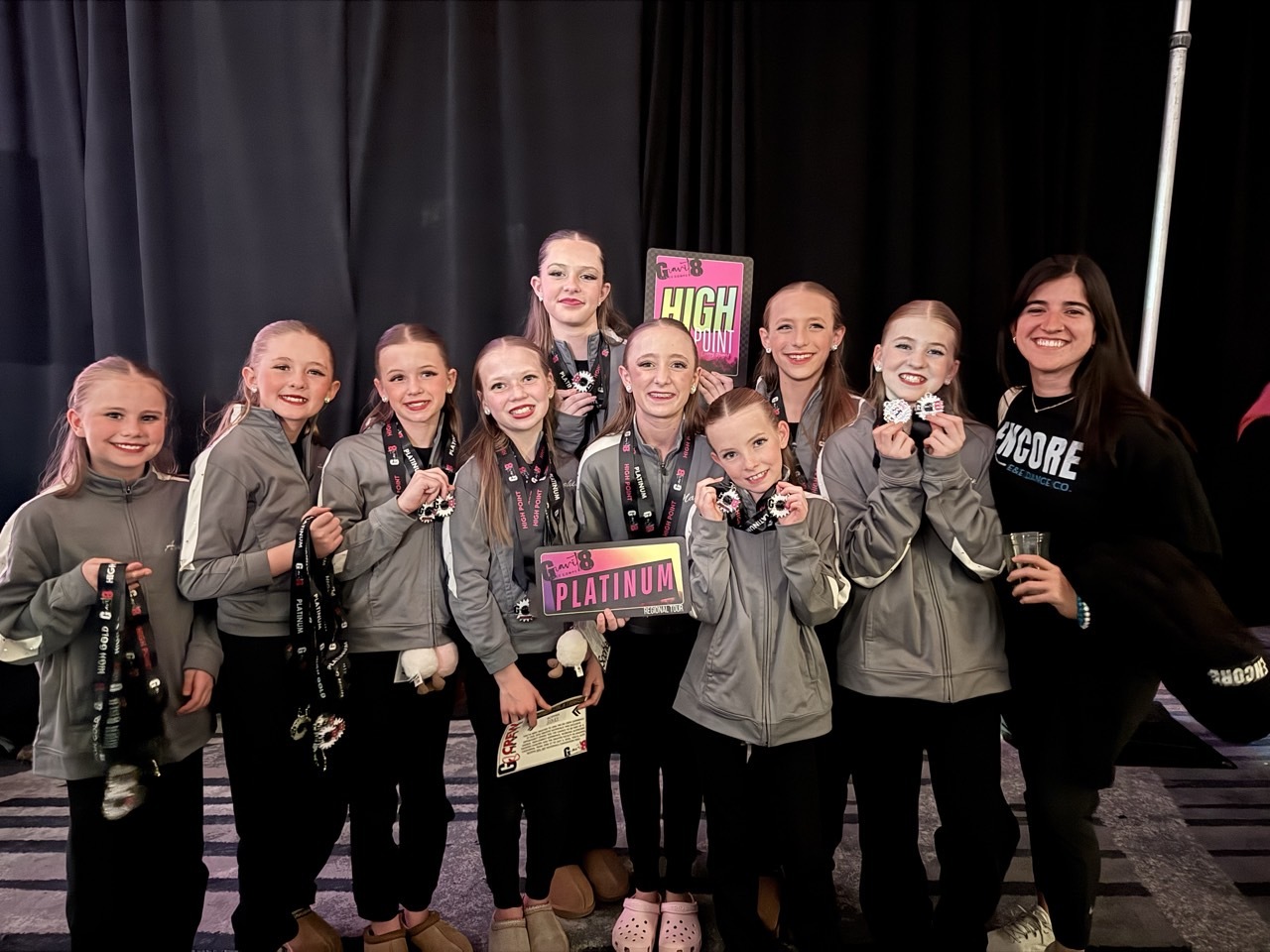 Encore dancers in matching gray jackets and their teacher smiling together with medals and competition awards.