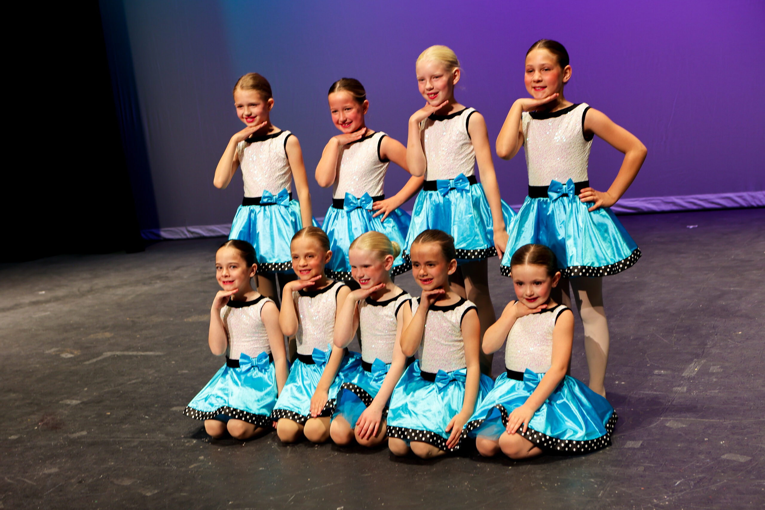 Nine young E&E Dance Company dancers posing on stage in white tops and turquoise skirts.