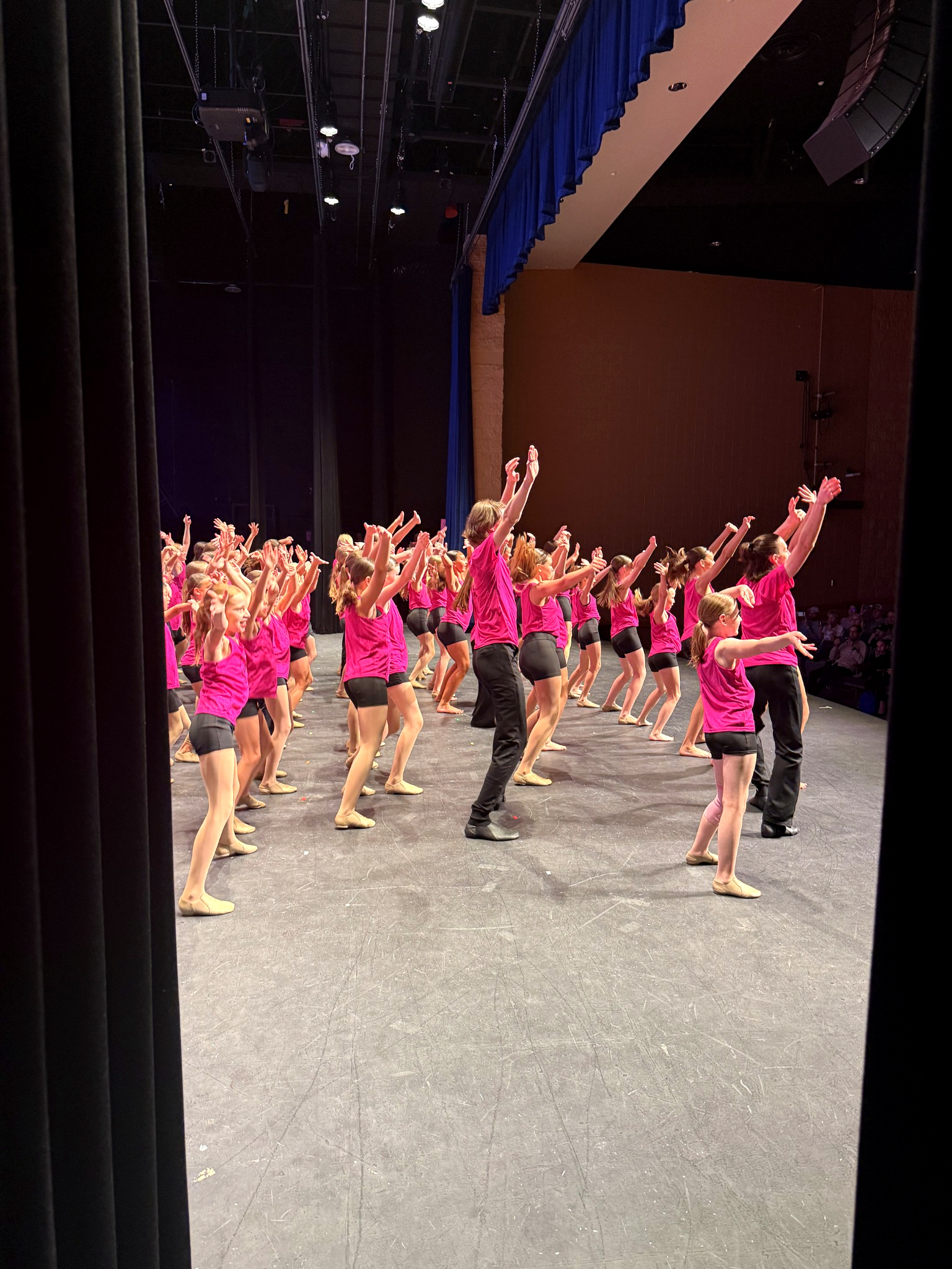 E&E Dance Company dancers in pink shirts performing together on stage with their arms raised.