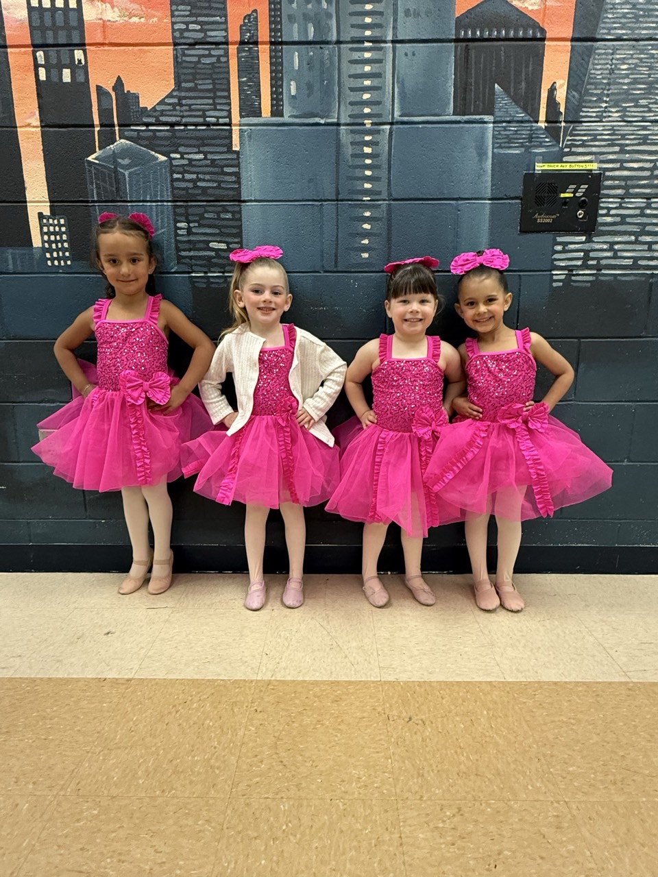 Four young dancers in bright pink recital costumes posing together in front of a city mural.
